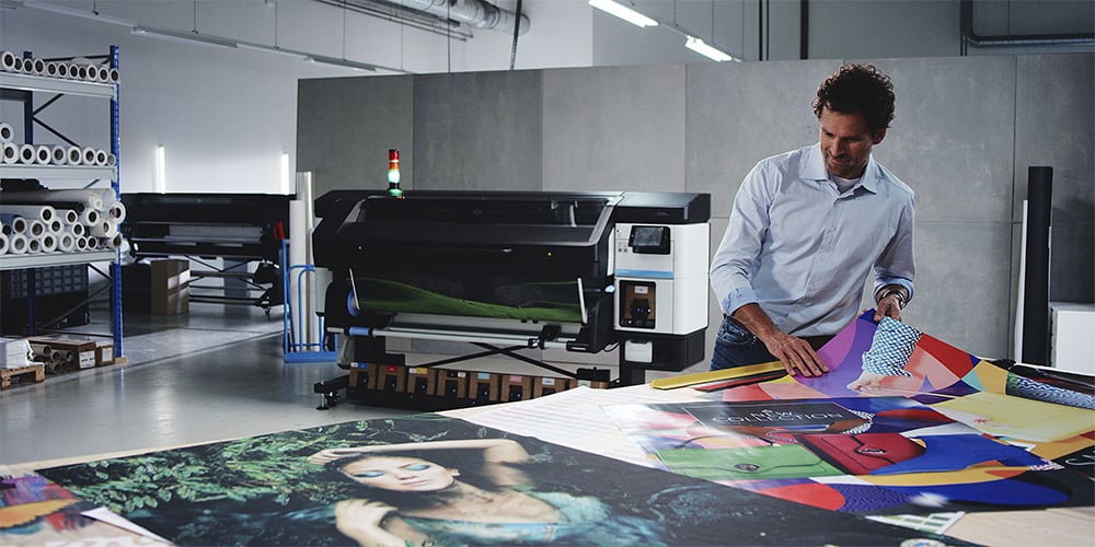 A person in a professional print workshop is laying out A0 posters on a table in front of a HP large-format printer