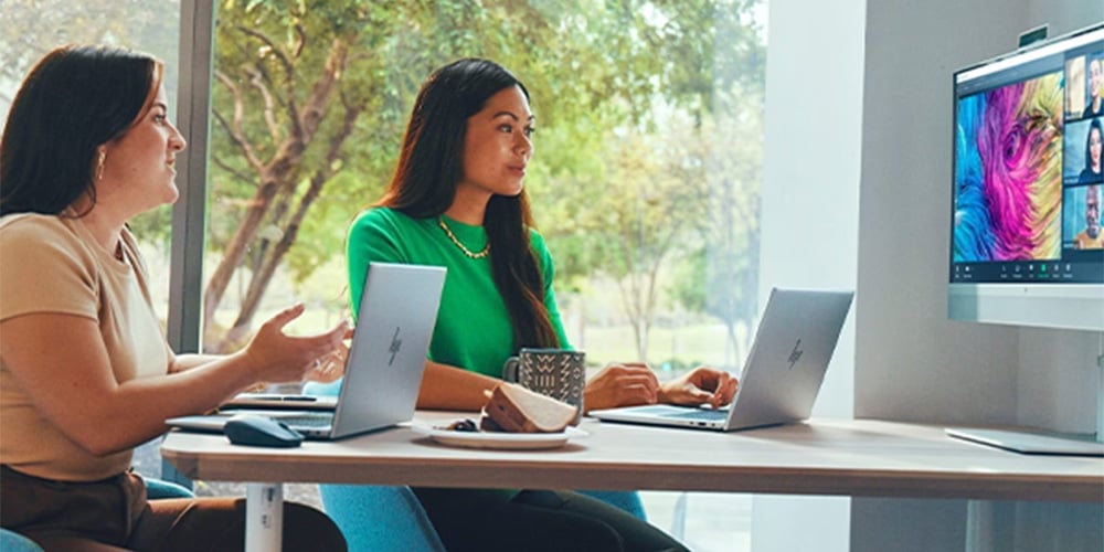 Two people working on their own HP laptops in an office setting, both facing a HP screen during a conference meeting.