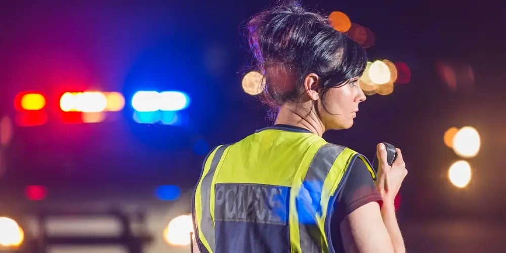 A police officer using a radio with emergency vehicle lights in the background.
