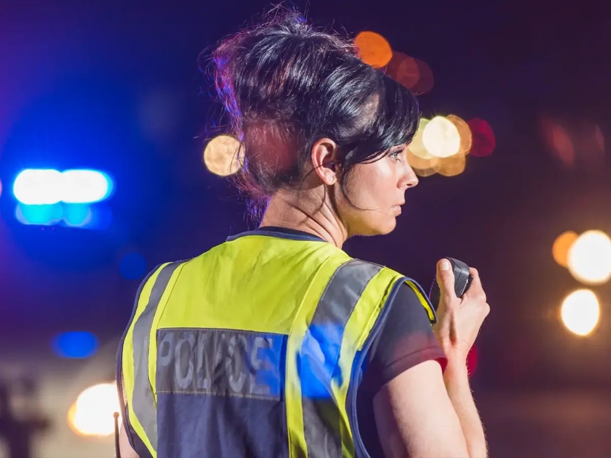 A police officer using a radio with emergency vehicle lights in the background.