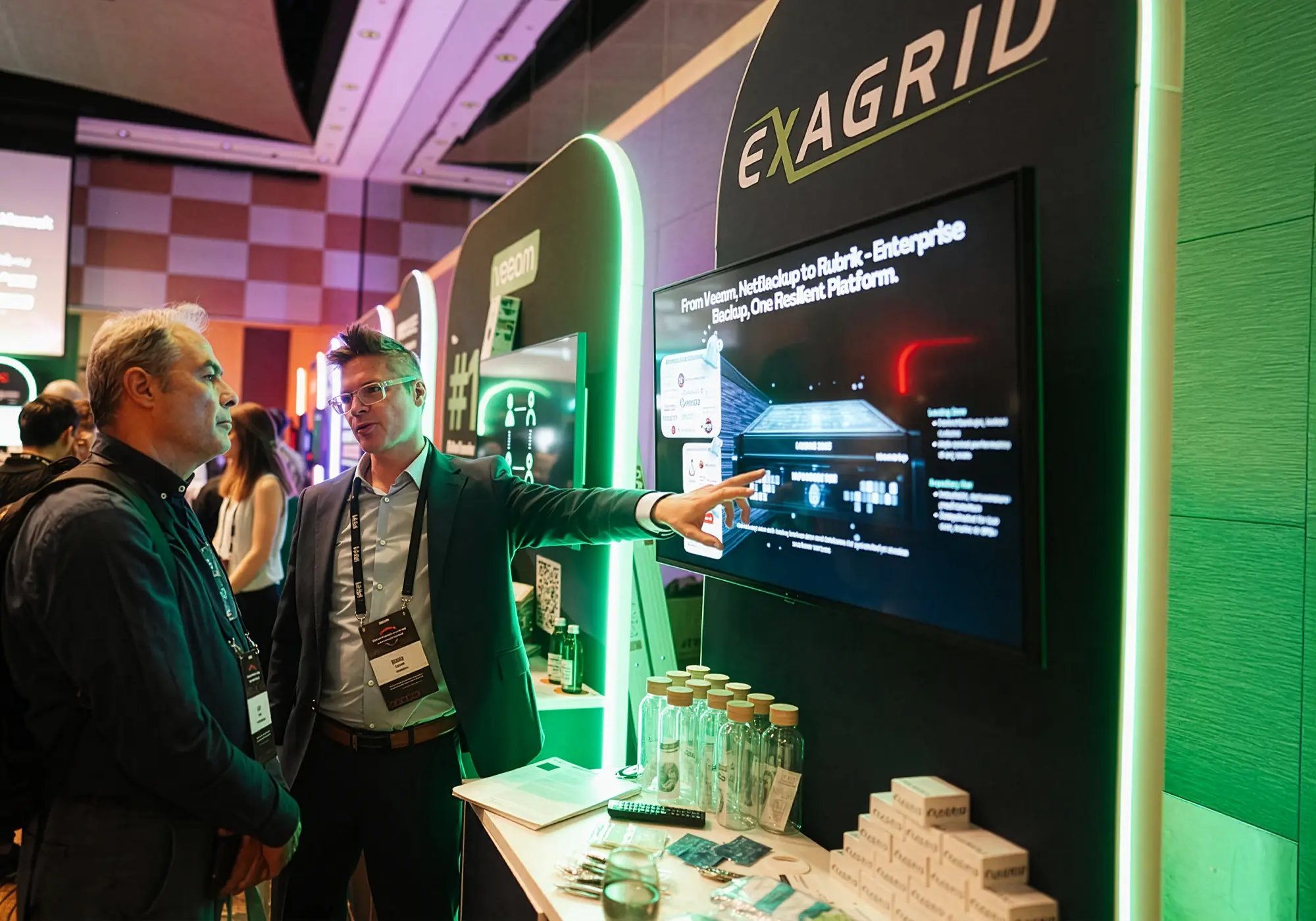 Man at tech conference booth gestures to digital display while explaining to another attendee. Booth features 