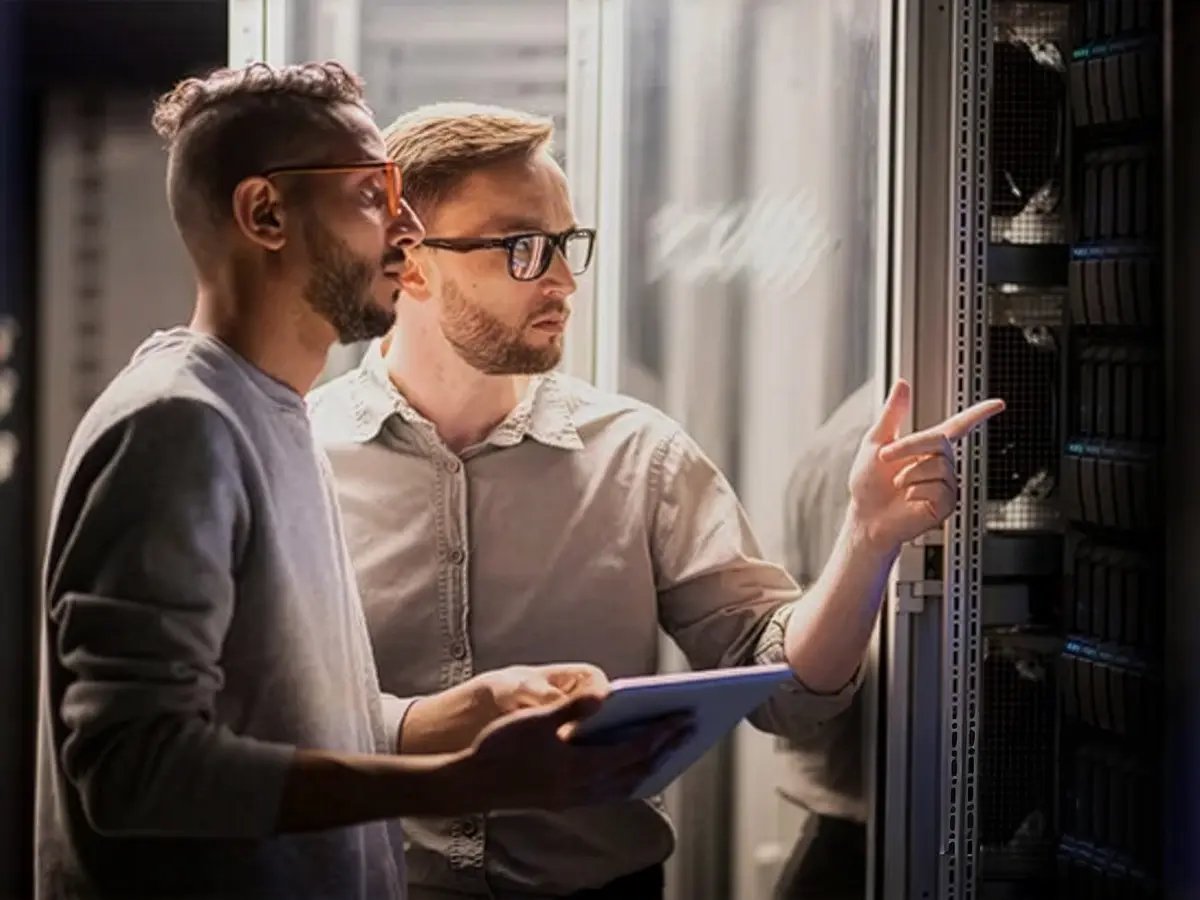 Two men in a server room, one pointing at a server rack, the other holding a tablet. They appear focused, suggesting a troubleshooting discussion.