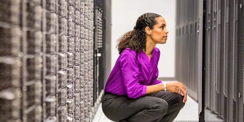 IT professional crouching in a data centre aisle, inspecting server racks and cabling between equipment rows.