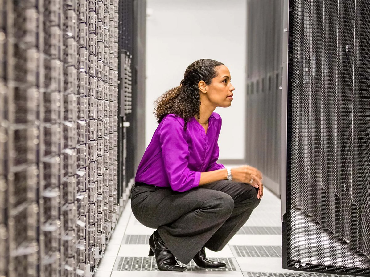 IT professional crouching in a data centre aisle, inspecting server racks and cabling between equipment rows.