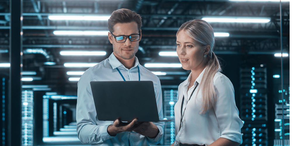 Two professionals stand in a server room, examining a laptop. The surroundings are filled with glowing servers, conveying a high-tech and focused atmosphere.