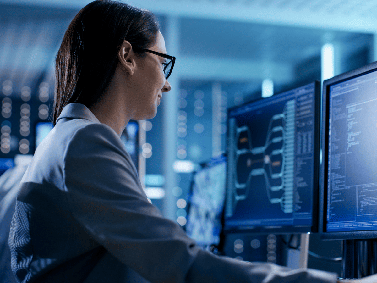 A woman in glasses and a blue blazer works at computer monitors displaying complex data and graphs in a dimly lit, modern office. The scene conveys focus.