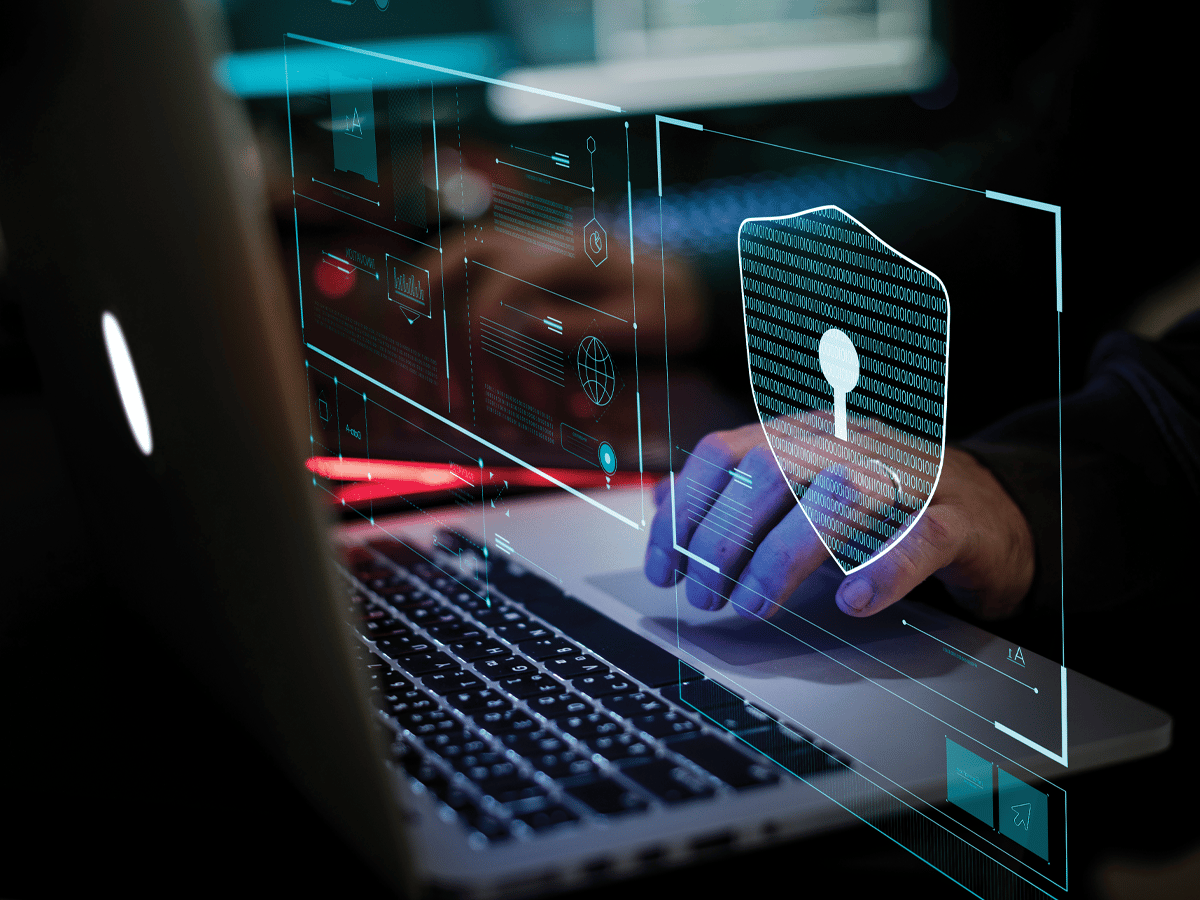 Close-up of hands using a laptop keyboard with a digital cybersecurity interface overlay, including a shield icon, binary code, and virtual security panels representing data protection and network security.