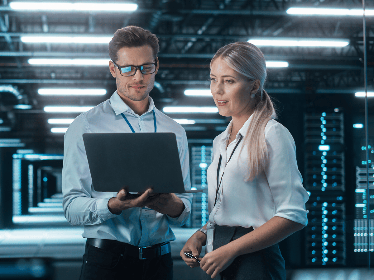 Two professionals stand in a server room, examining a laptop. The surroundings are filled with glowing servers, conveying a high-tech and focused atmosphere.