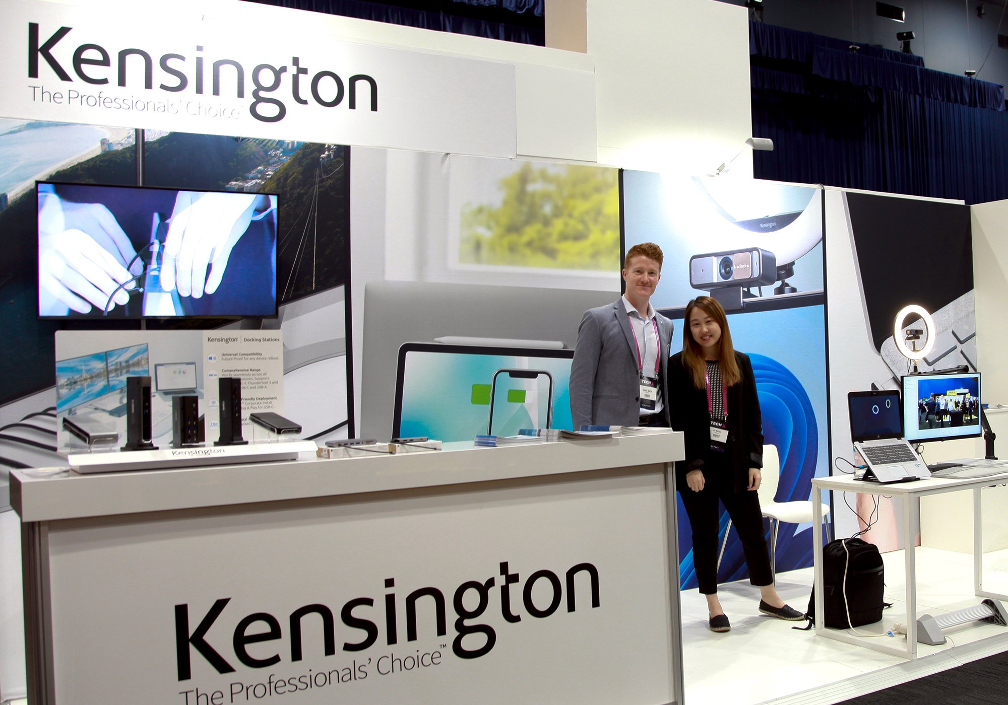 A trade show booth displaying Kensington products, with two people standing near a counter featuring tech accessories and promotional screens