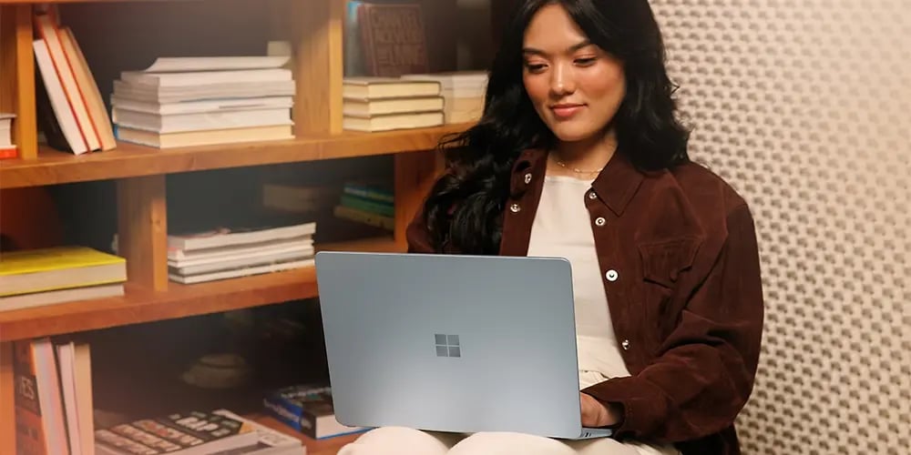 A person working on a Microsoft Surface laptop while seated in front of a bookshelf.