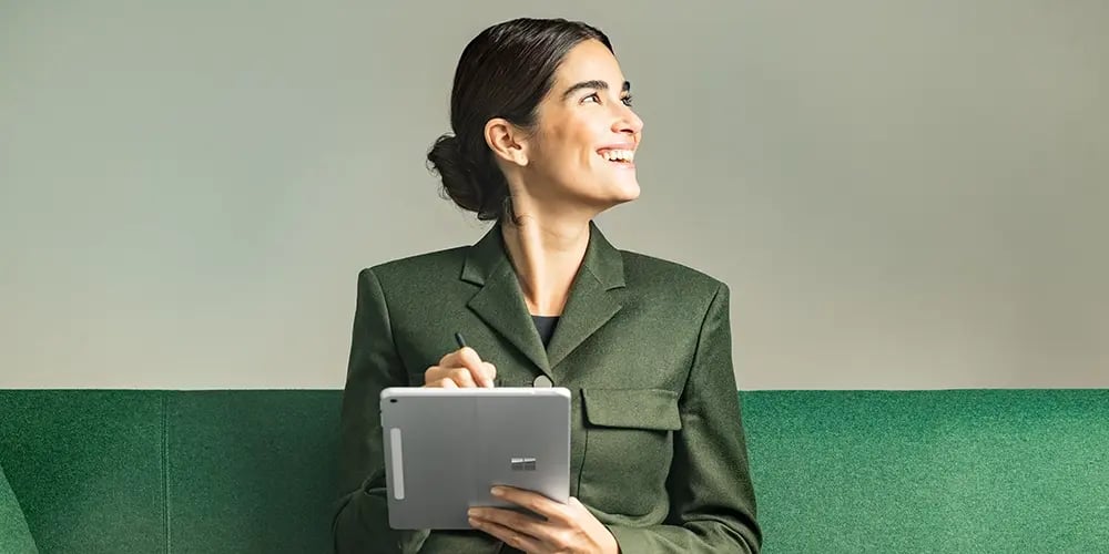 A person holding a Surface tablet and pen while seated on a green bench.