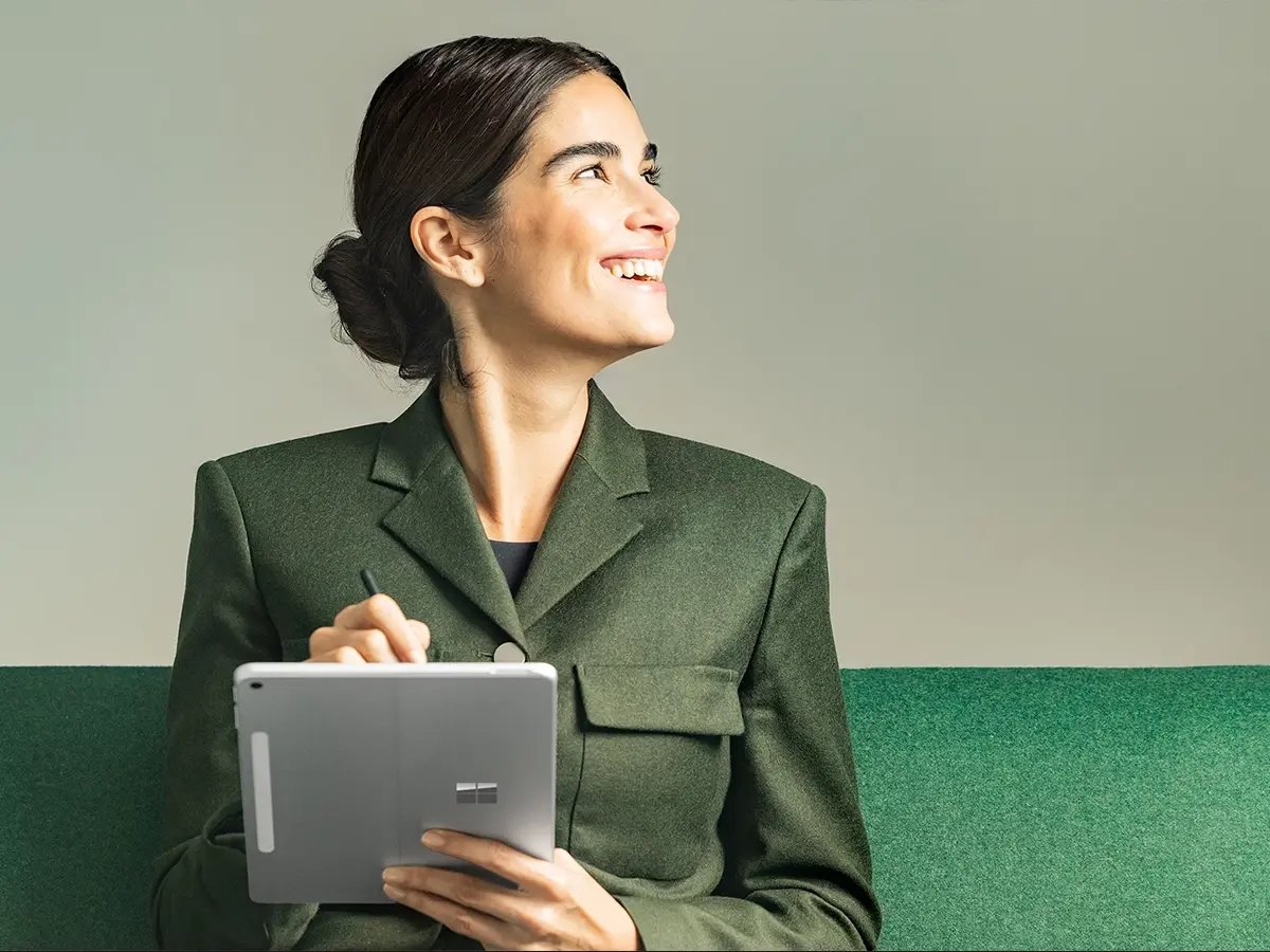 A person holding a Surface tablet and pen while seated on a green bench.
