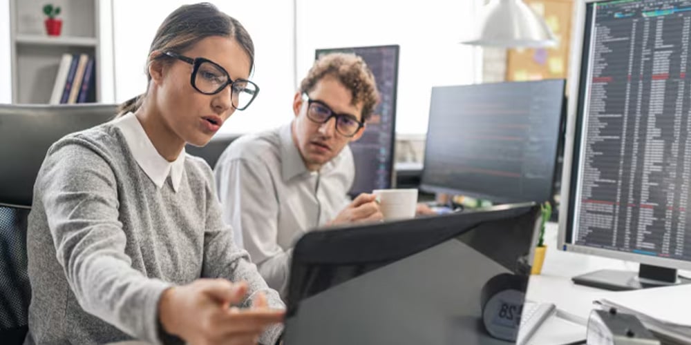 Two colleagues collaborating at a desk, reviewing information on a large monitor in an office.