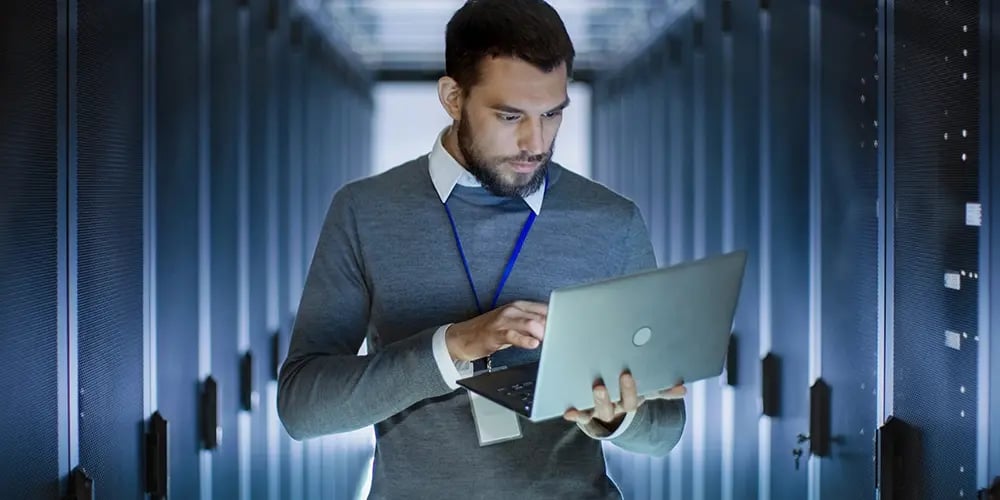 A person using a laptop while standing in a data‑center aisle.