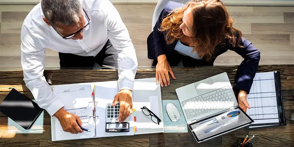 Two people working at a desk with documents, a calculator, and a laptop.