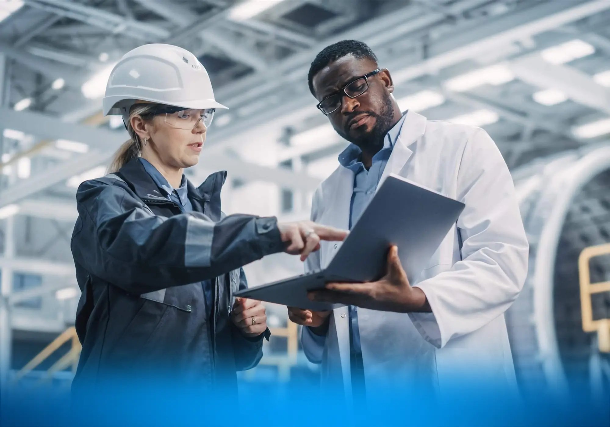 Two workers in safety gear reviewing information on a laptop inside an industrial facility.