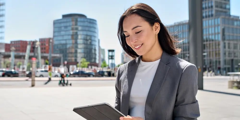 A woman in a gray blazer smiles while using a tablet outdoors in a modern urban setting. Tall glass buildings and parked cars are visible in the background.