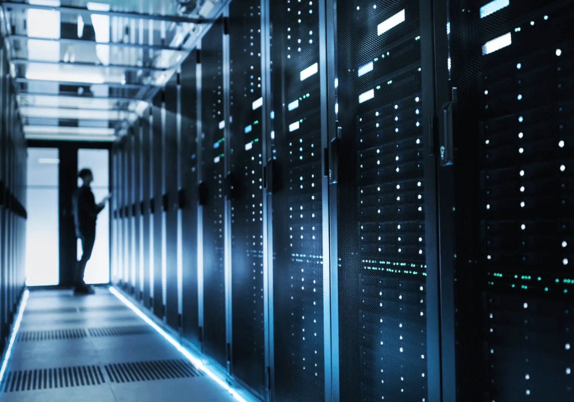 A person stands in a dimly lit server room, illuminated by blue lights. Tall server racks line the corridor, creating a high-tech and futuristic atmosphere.