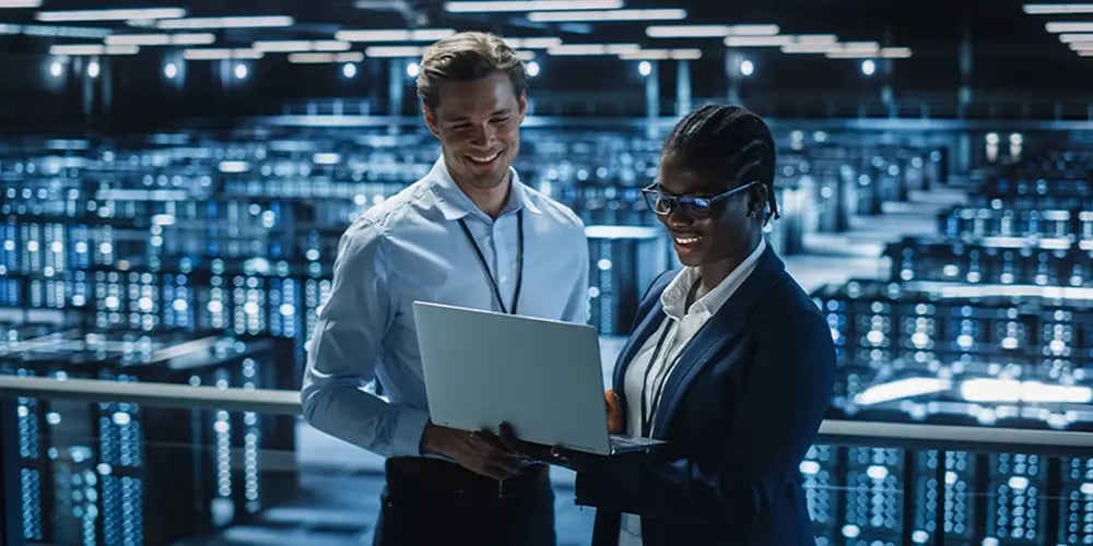 Two professionals in business attire view a laptop in a large, brightly lit data center. They appear engaged and smiling, suggesting a positive atmosphere.