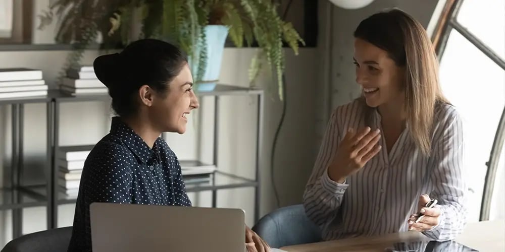 Two women are sitting at a wooden table, engaged in a friendly conversation. One holds a phone, and the other has a laptop. They're smiling warmly.
