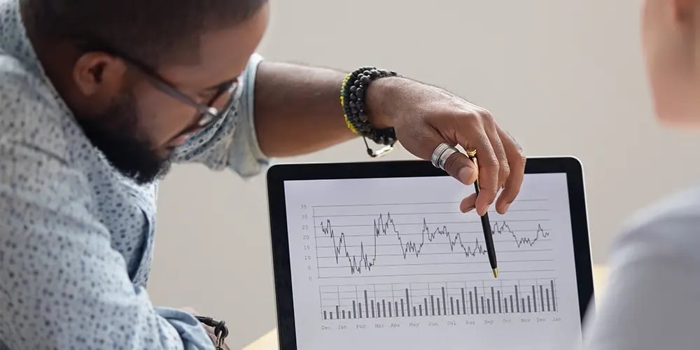 A person points at a line graph and bar chart on a tablet during a discussion. The graphs show fluctuating data trends, indicating analysis or strategic planning.