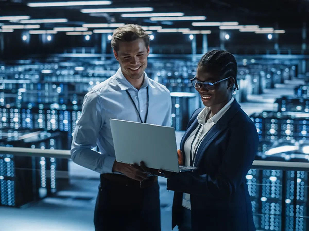 Two professionals in business attire view a laptop in a large, brightly lit data center. They appear engaged and smiling, suggesting a positive atmosphere.