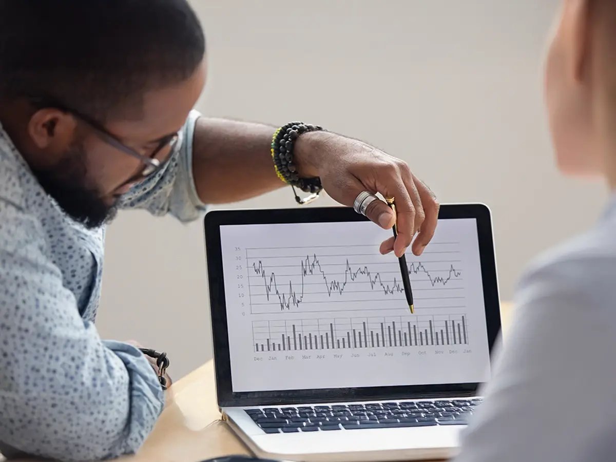 A person points at a line graph and bar chart on a tablet during a discussion. The graphs show fluctuating data trends, indicating analysis or strategic planning.