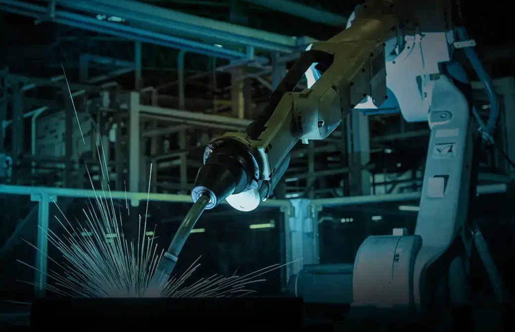 Industrial robotic arm welding metal with bright sparks in a factory setting.