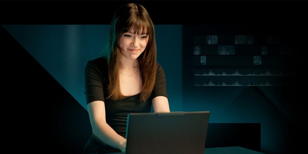Person typing on a laptop in a dark room with data charts faintly visible on the wall behind.