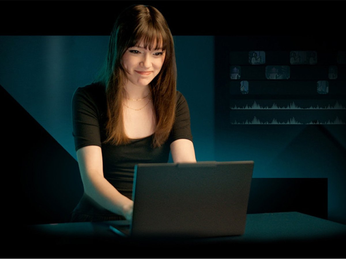 Person typing on a laptop in a dark room with data charts faintly visible on the wall behind.