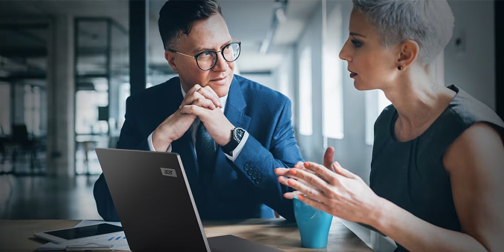 Two people sitting at a desk in an office, discussing work with an open laptop and coffee cup.
