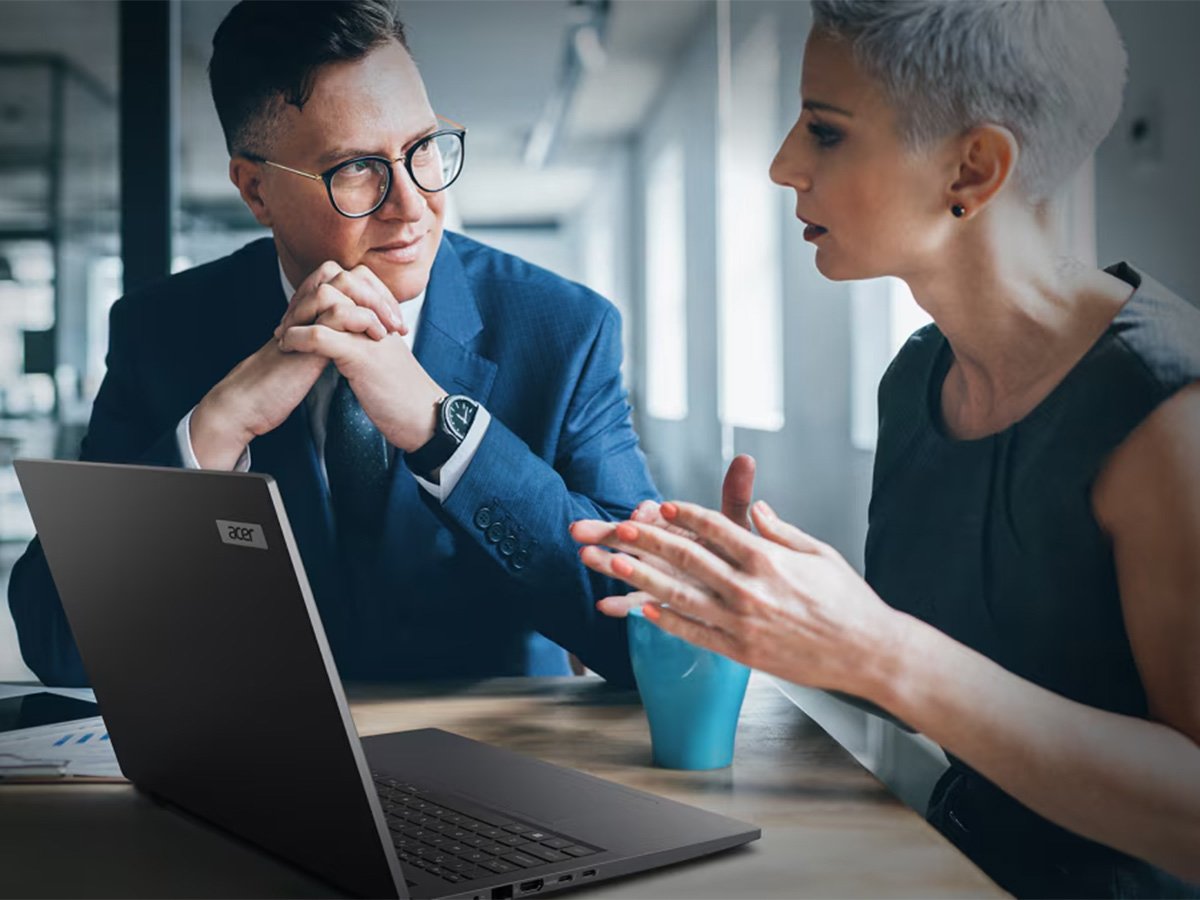 Two people sitting at a desk in an office, discussing work with an open laptop and coffee cup.