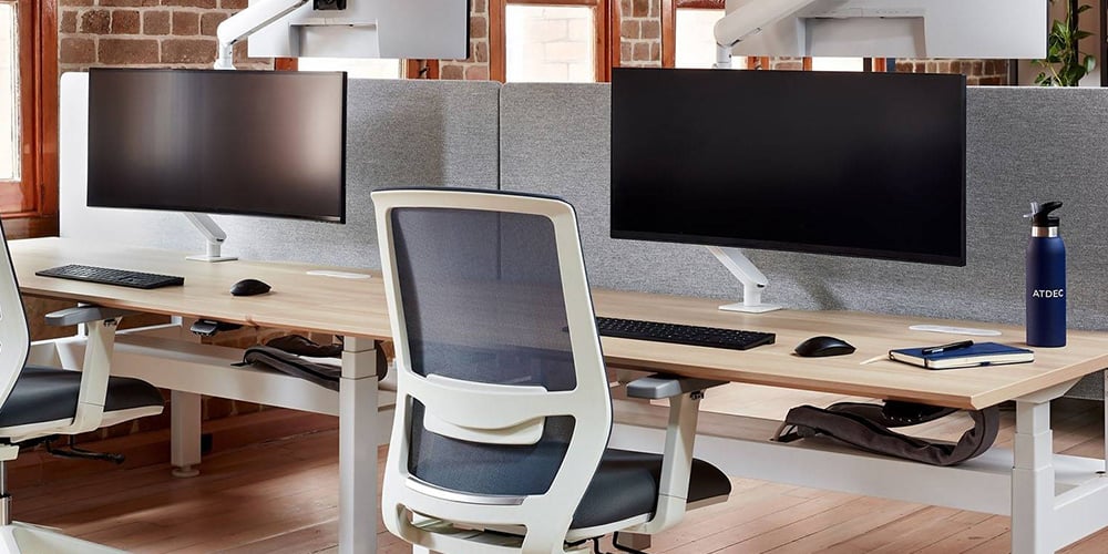 Modern office setup with dual monitor arms, ergonomic chairs, and wooden desks near large windows.