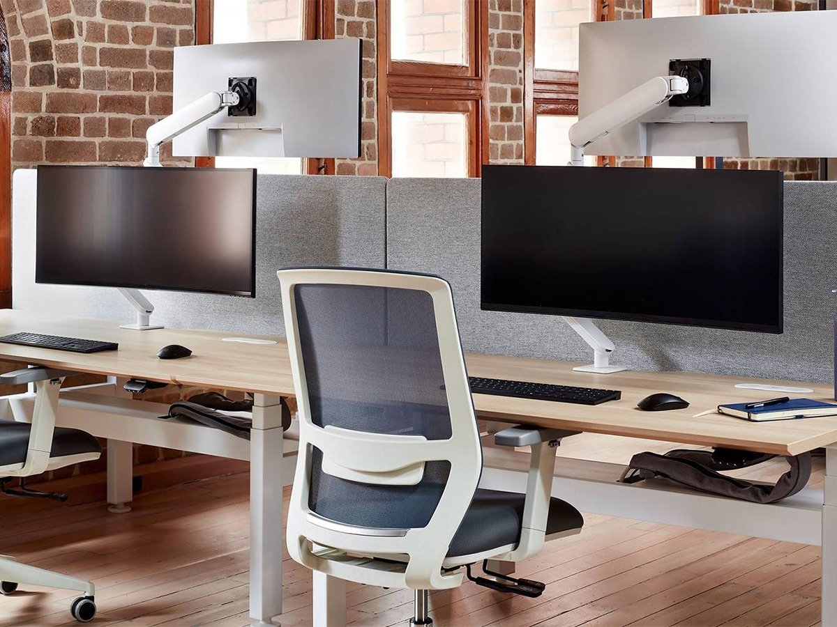Modern office setup with dual monitor arms, ergonomic chairs, and wooden desks near large windows.