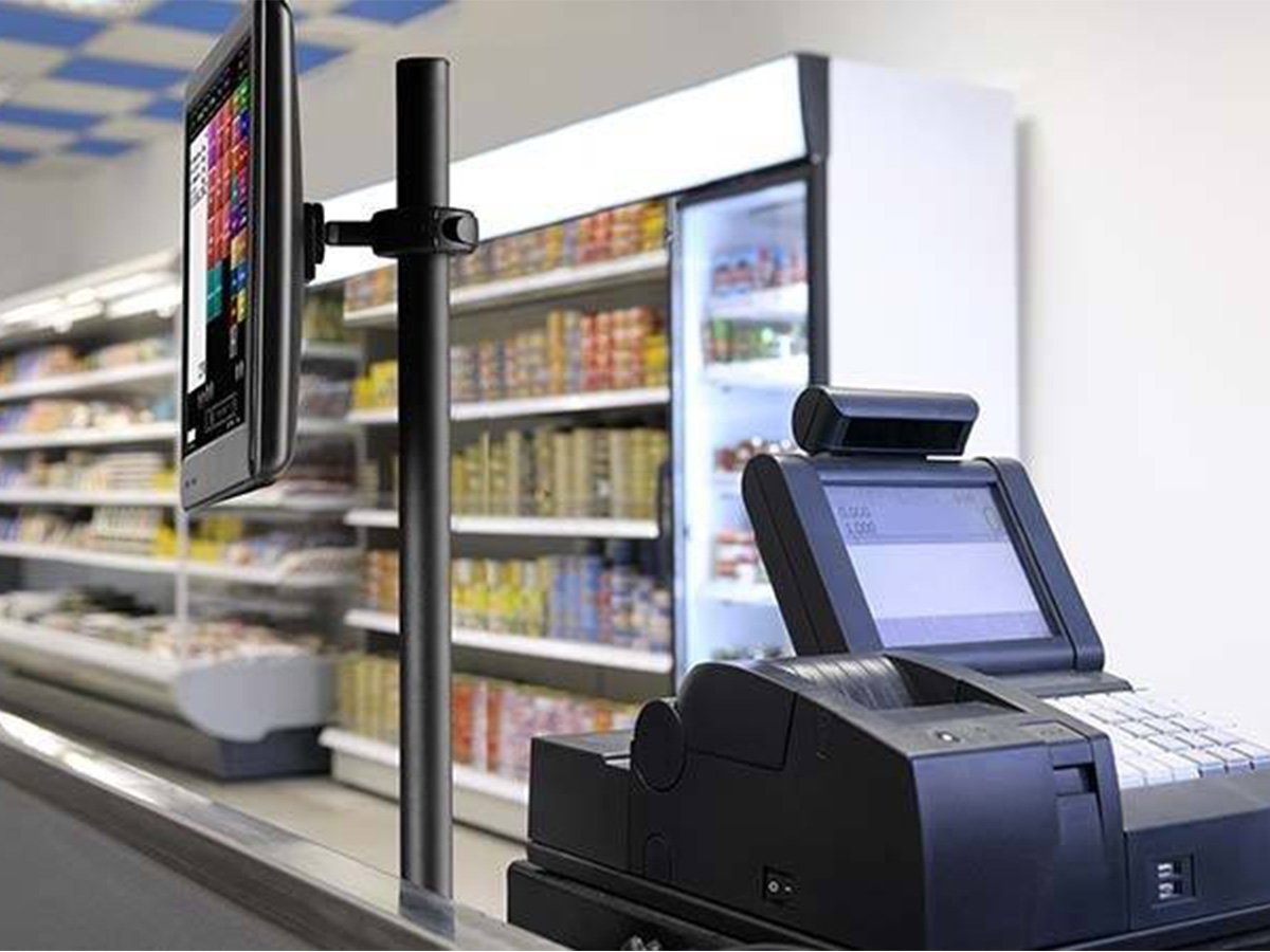 Retail checkout counter with a touchscreen monitor and payment terminal in front of stocked shelves.