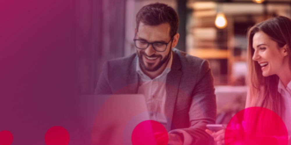 Two people sitting at a table in a modern office, working on a laptop with pink graphic overlays.