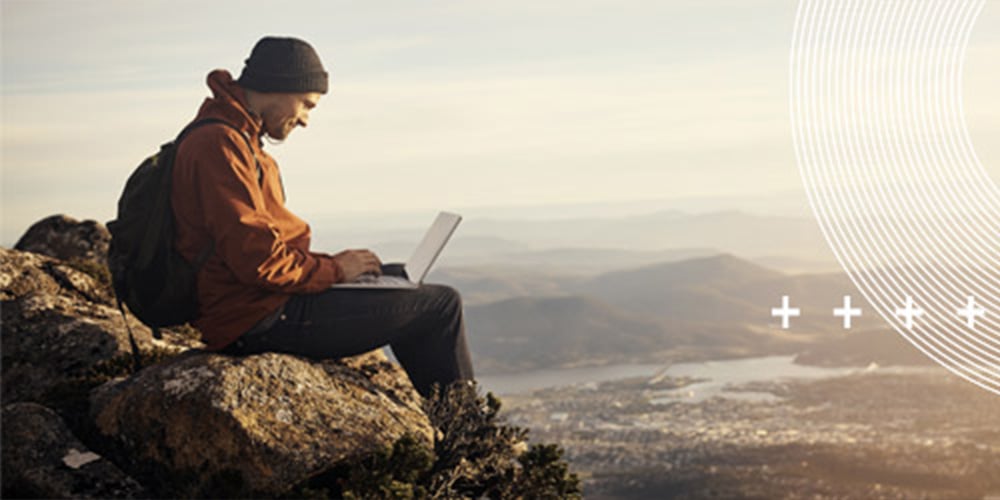 Person sitting outdoors on a rocky hilltop using a laptop with mountains in the background.