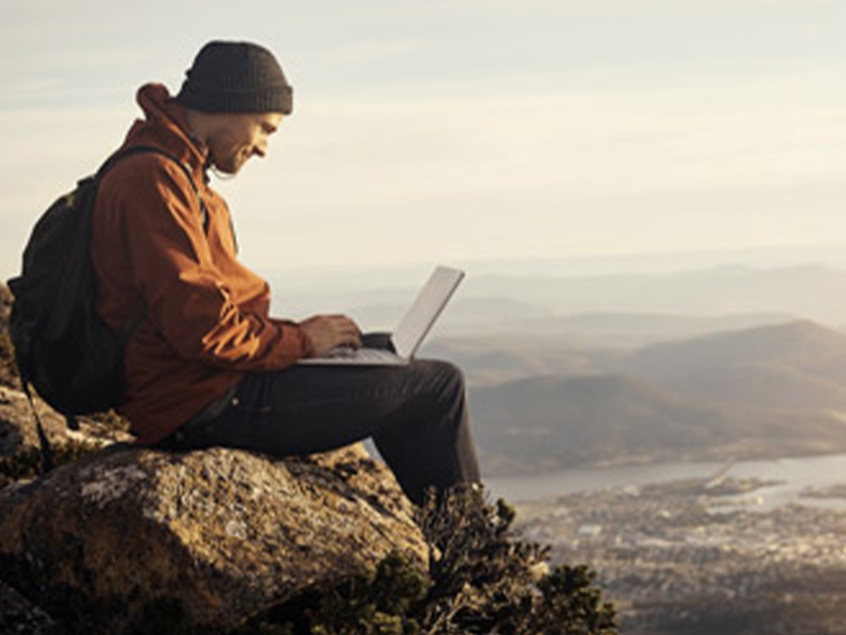 Person sitting outdoors on a rocky hilltop using a laptop with mountains in the background.