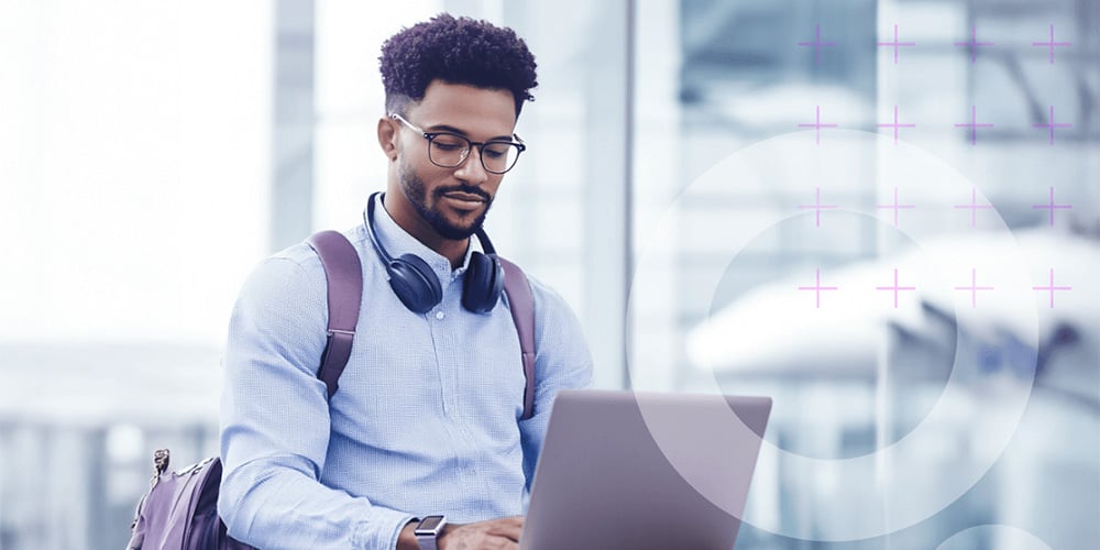 Person seated in an airport terminal using a laptop with headphones around the neck and a bag beside.