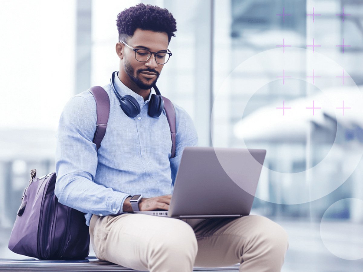 Person seated in an airport terminal using a laptop with headphones around the neck and a bag beside.