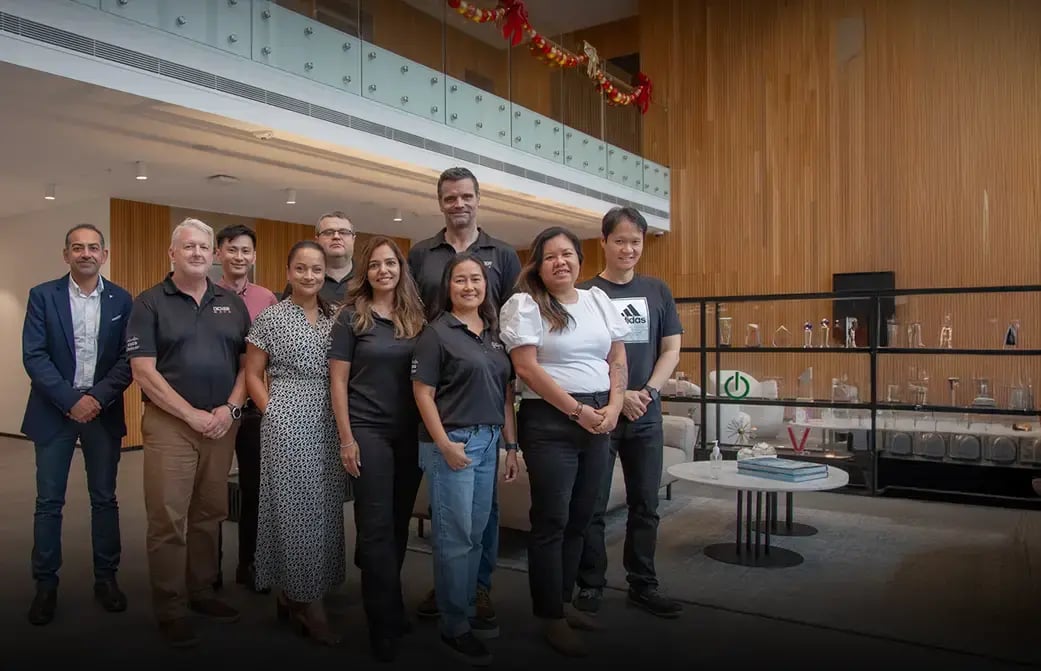 Group of people standing in a modern office lobby with wooden walls, glass railing, and award display shelf.