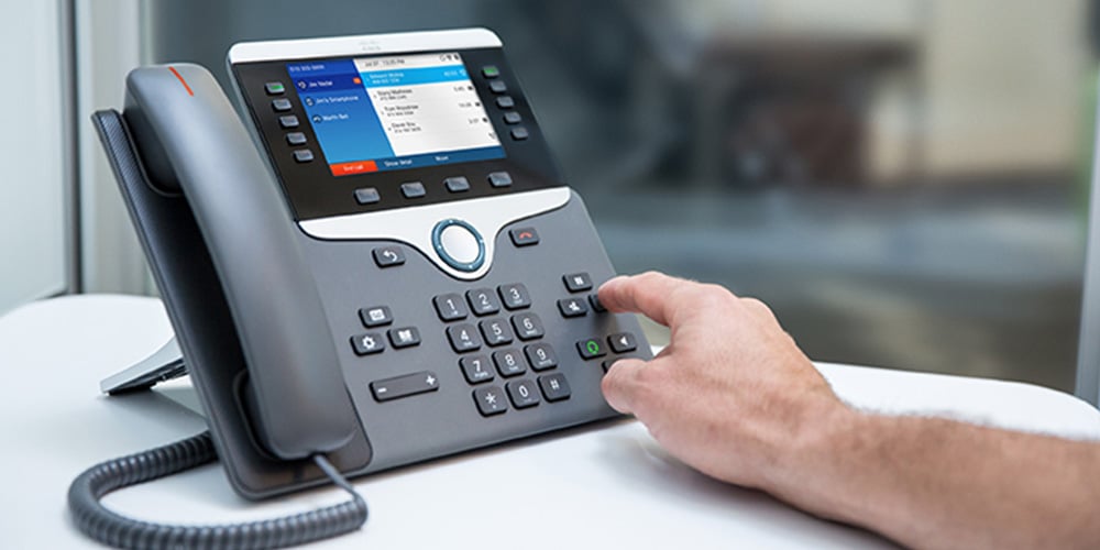 Person pressing buttons on a Cisco office desk phone with a digital display showing call options.