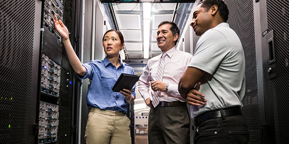 Three people in a server room with racks of equipment, one pointing at servers while holding a tablet.