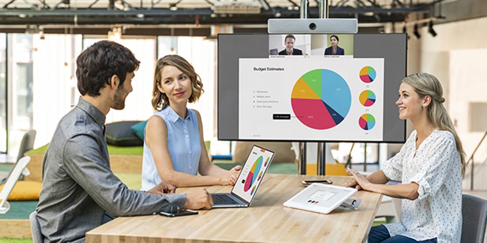 People in a meeting room with a video conference screen showing colorful charts and remote participants.