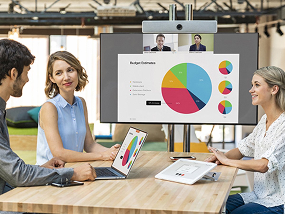 People in a meeting room with a video conference screen showing colorful charts and remote participants.