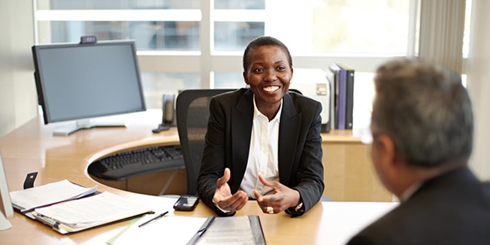 Two people sitting at a desk in an office, talking with documents and a smartphone on the table.