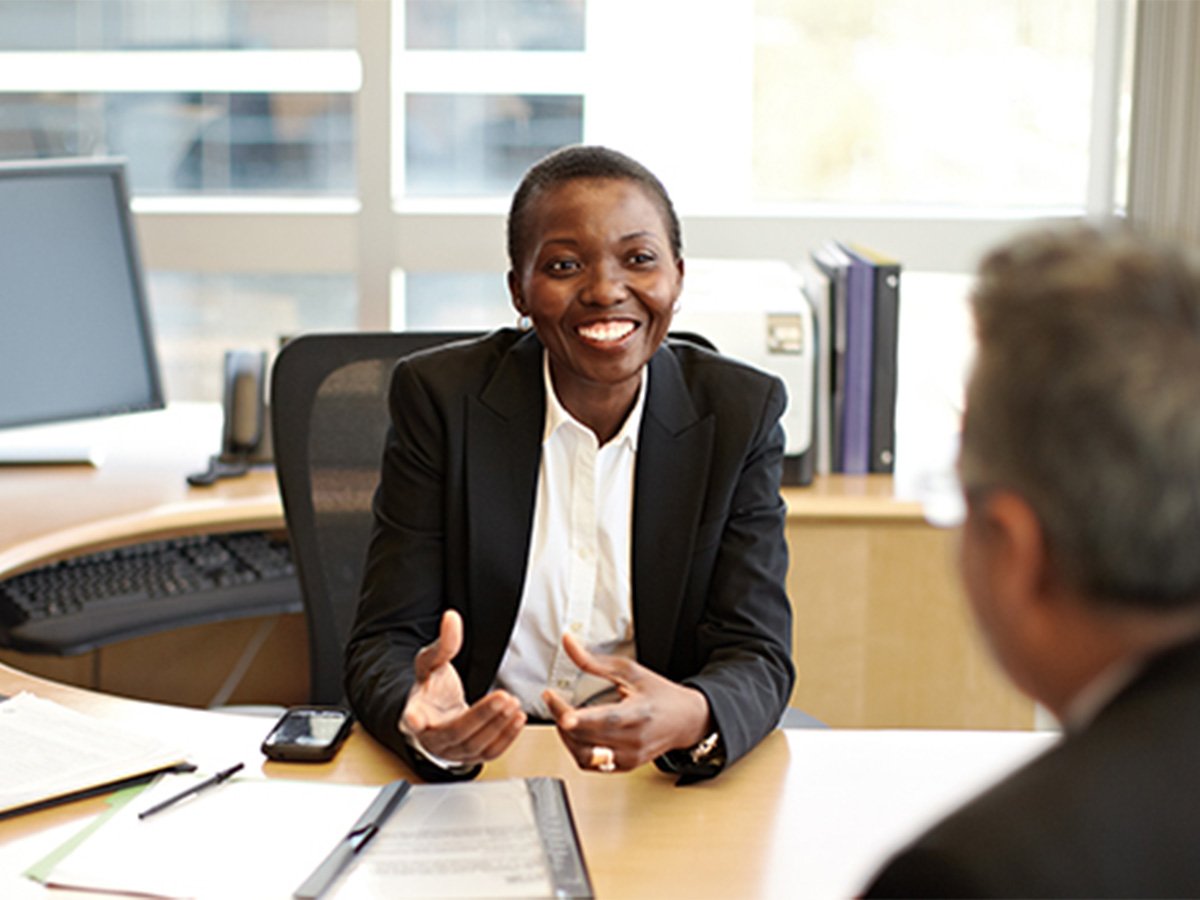 Two people sitting at a desk in an office, talking with documents and a smartphone on the table.