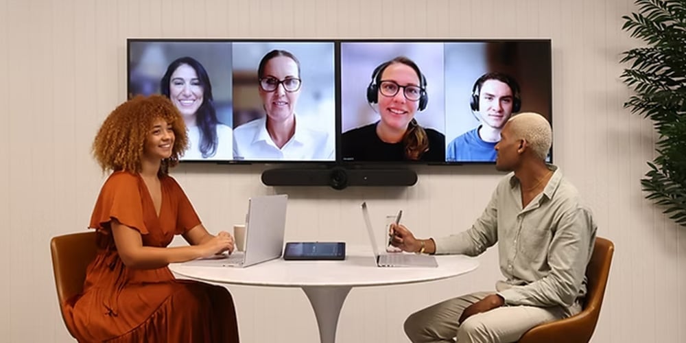 Two people seated at a table using laptops during a video conference on a wall-mounted screen.