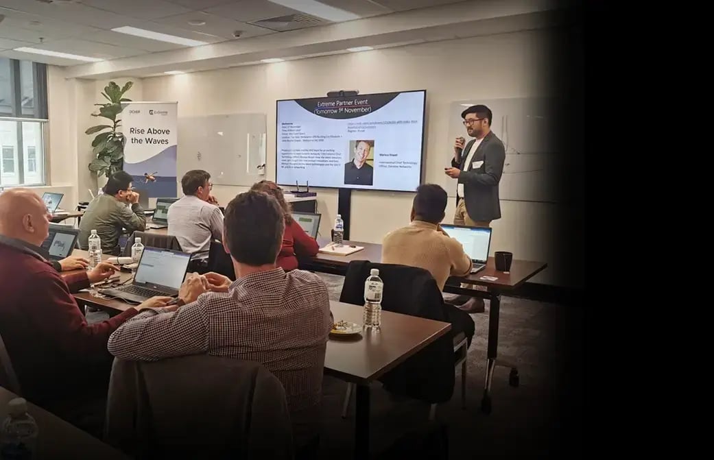 Presenter speaking to attendees in a classroom setup, with laptops open and a slide displayed on a large