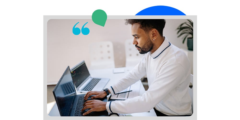 Person working on a laptop at a desk with a second laptop and workspace items nearby.